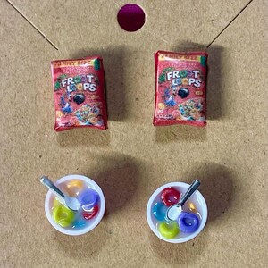 May include: A pair of earrings featuring miniature cereal boxes and bowls of cereal. The cereal boxes are red and say "Frosted Flakes" on them. The bowls are white with a silver spoon and colorful cereal inside.