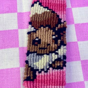 May include: A pink and white woven friendship bracelet with a brown and white pixelated image of a cartoon character.