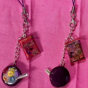 May include: Two phone charms featuring a cereal box and a bowl of cereal. The cereal box charm is red and features the text "Frosted Flakes". The bowl of cereal charm is black and features a white bowl with colorful cereal and milk.