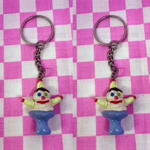 May include: Two ice cream cone keychain charms with a smiling face, green and blue eyes, and a pink mouth. The cones are yellow and white with a brown swirl on top. The cones are on a blue stand and attached to silver keychains.