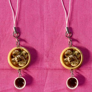 May include: Two white string charm necklaces with a small yellow bowl filled with dumplings and a small cup of sauce. The necklaces are on a pink fabric background.