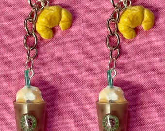 May include: Two keychains with a brown iced coffee charm and a yellow croissant charm. The charms are attached to silver keychains.