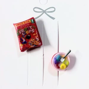 May include: A pair of miniature cereal-themed earrings. The first earring is a red box of Froot Loops cereal. The second earring is a pink bowl of cereal with a silver spoon.