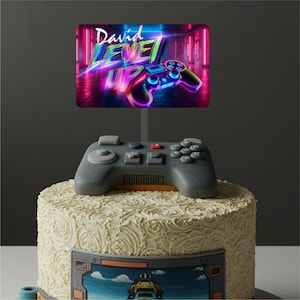 May include: A white cake with a grey video game controller on top. The cake is decorated with a white frosting that looks like swirls. There is a cake topper that says "David Level Up" with a neon pink and blue video game controller on it.
