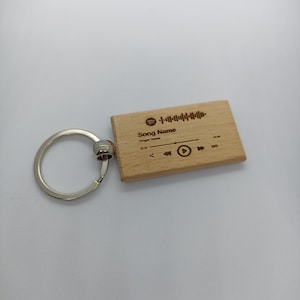 Op de afbeelding: Een houten sleutelhanger met een muziekspelerontwerp. De sleutelhanger heeft een afspeelknop, een pauzeknop en een skipknop. De tekst "Song Name" is gegraveerd op de sleutelhanger.