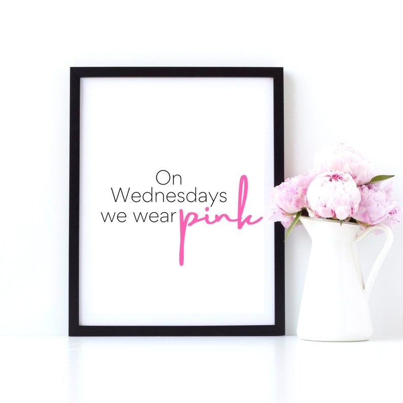May include: A black framed print with the text "On Wednesdays we wear pink" in white and pink lettering. The print is sitting on a white surface next to a white pitcher with pink flowers.