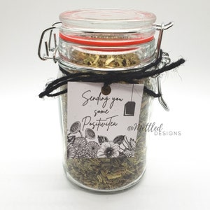 May include: A clear glass jar filled with loose leaf tea, sealed with a red-trimmed lid and metal clasp. A black string is tied around the jar, holding a tag that reads "Sending you some PositiviTea" with floral illustrations.