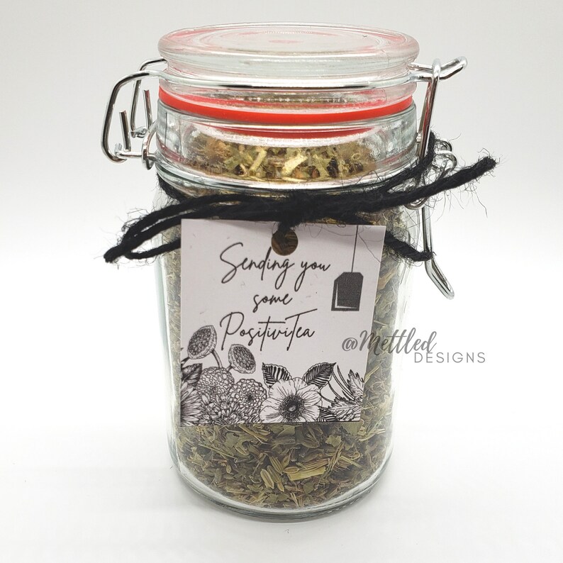 May include: A clear glass jar filled with loose leaf tea, sealed with a red-trimmed lid and metal clasp. A black string is tied around the jar, holding a tag that reads "Sending you some PositiviTea" with floral illustrations.