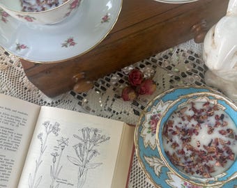 May include: A vintage teacup and saucer set filled with a creamy liquid and dried rose petals. The teacup and saucer have a floral design with gold trim. An open book with botanical illustrations and a decorative ceramic bunny are also visible.
