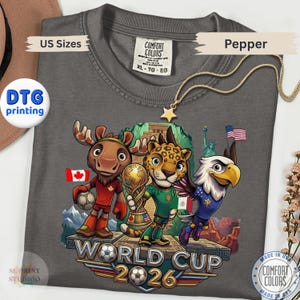 May include: A charcoal gray t-shirt featuring a graphic of a cartoon moose, jaguar, and bald eagle standing on a platform. The characters hold a soccer ball and a trophy. The text reads World Cup 2026. The shirt includes a Comfort Colors brand label.