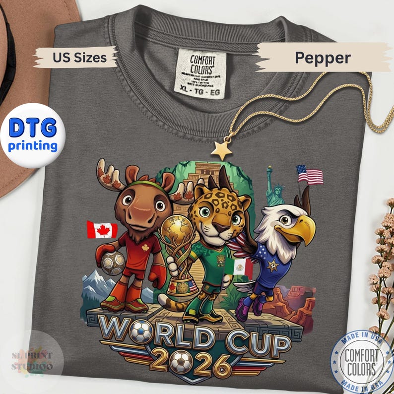 May include: A charcoal gray t-shirt featuring a graphic of a cartoon moose, jaguar, and bald eagle standing on a platform. The characters hold a soccer ball and a trophy. The text reads World Cup 2026. The shirt includes a Comfort Colors brand label.