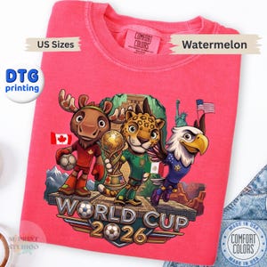 May include: A watermelon-colored crewneck sweatshirt featuring a graphic of a cartoon moose, jaguar, and bald eagle standing on a stone platform. The design includes a soccer trophy, the text WORLD CUP 2026, and flags representing Canada, Mexico, and the USA.