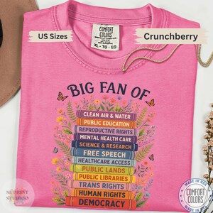 以下が含まれることがあります： BIG FAN OFという文字と本の積み重ねのグラフィックが描かれたピンクのTシャツ。本の背表紙にはCLEAN AIR & WATER、PUBLIC EDUCATION、REPRODUCTIVE RIGHTS、DEMOCRACYなどの社会的なテーマが記されており、周囲には花が描かれています。