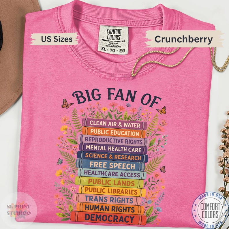 以下が含まれることがあります： BIG FAN OFという文字と本の積み重ねのグラフィックが描かれたピンクのTシャツ。本の背表紙にはCLEAN AIR & WATER、PUBLIC EDUCATION、REPRODUCTIVE RIGHTS、DEMOCRACYなどの社会的なテーマが記されており、周囲には花が描かれています。