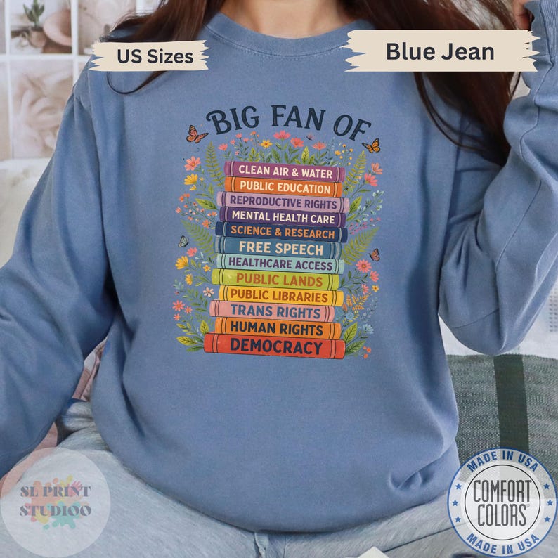 以下が含まれることがあります： ブルーデニムカラーのクルーネックトレーナー。中央には「BIG FAN OF」という文字と、カラフルな本の積み重ねのグラフィックが描かれています。本の背表紙には、Clean Air & Water、Public Education、Reproductive Rights、Mental Health Care、Science & Research、Free Speech、Healthcare Access、Public Lands、Public Libraries、Trans Rights、Human Rights、Democracyと記されており、花や蝶のイラストが添えられています。