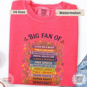 以下が含まれることがあります： スイカ色のクルーネックTシャツ。花と蝶に囲まれた本の積み重ねのグラフィックが描かれています。本の背表紙には「BIG FAN OF」という文字と、様々な社会問題がリスト形式でプリントされています。
