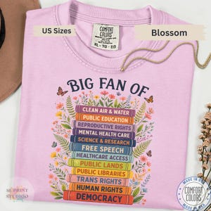 以下が含まれることがあります： 本が積み重なったグラフィックと「BIG FAN OF」というテキスト、そしてClean Air & Water、Public Education、Reproductive Rights、Mental Health Care、Science & Research、Democracyなどの社会問題がリストされた薄いピンクのTシャツ。