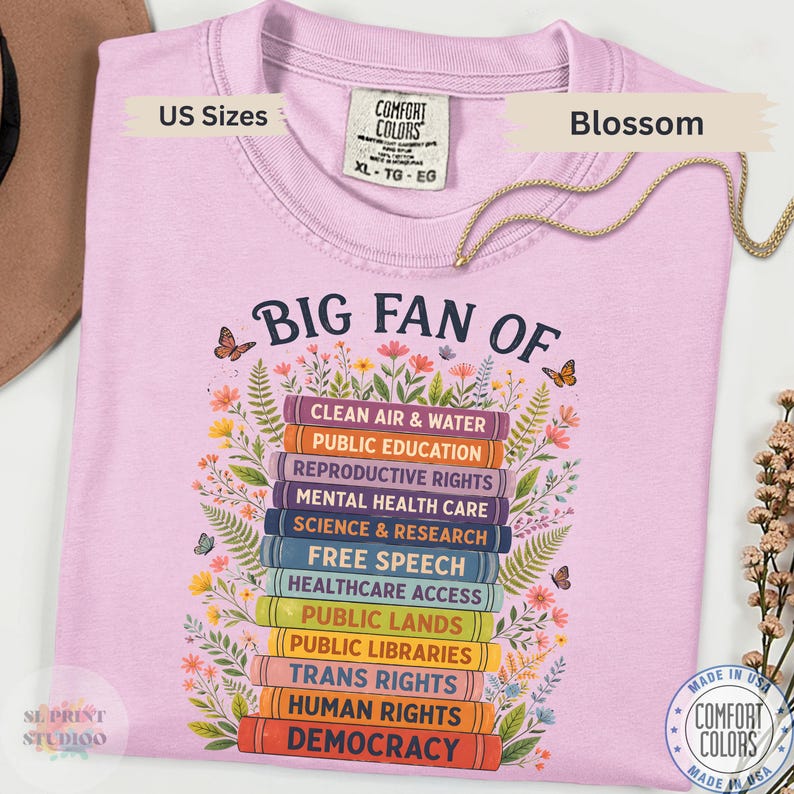 以下が含まれることがあります： 本が積み重なったグラフィックと「BIG FAN OF」というテキスト、そしてClean Air & Water、Public Education、Reproductive Rights、Mental Health Care、Science & Research、Democracyなどの社会問題がリストされた薄いピンクのTシャツ。