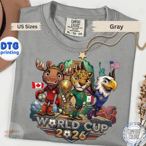 May include: A gray t-shirt featuring a graphic of a cartoon moose, jaguar, and bald eagle standing on a platform. The moose holds a soccer ball, the jaguar holds a trophy, and the eagle flies nearby. Text reads World Cup 2026 with flags.