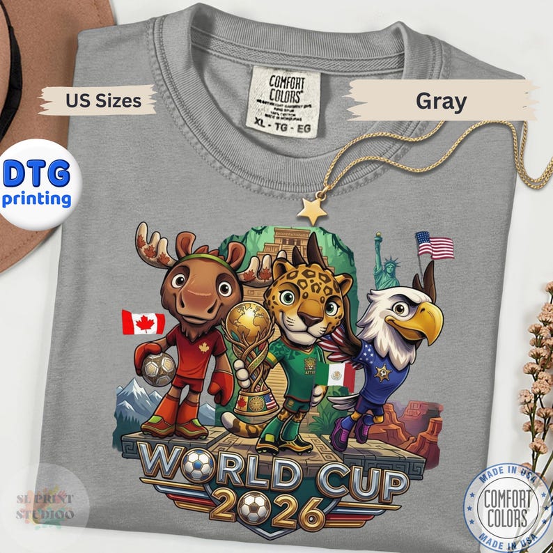 May include: A gray t-shirt featuring a graphic of a cartoon moose, jaguar, and bald eagle standing on a platform. The moose holds a soccer ball, the jaguar holds a trophy, and the eagle flies nearby. Text reads World Cup 2026 with flags.