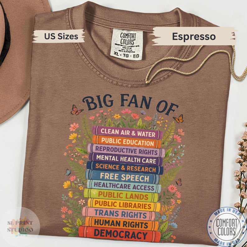 以下が含まれることがあります： エスプレッソカラーのTシャツで、花と蝶に囲まれたカラフルな本の積み重ねのグラフィックが特徴です。テキストには「BIG FAN OF」とあり、その後にClean Air & Water、Public Education、Reproductive Rights、Mental Health Care、Science & Research、Free Speech、Healthcare Access、Public Lands、Public Libraries、Trans Rights、Human Rights、Democracyと書かれています。