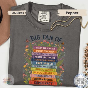 以下が含まれることがあります： チャコールグレーのTシャツで、色とりどりの花や蝶に囲まれた本の積み重ねのグラフィックが描かれています。テキストには「BIG FAN OF」とあり、その後に「Clean Air & Water」、「Public Education」、「Democracy」などの社会的な大義名分がリストされています。