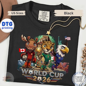 May include: A black t-shirt featuring a graphic of a cartoon moose, jaguar, and bald eagle standing on a platform with a soccer ball and trophy. The text reads World Cup 2026. The shirt includes a Comfort Colors label in size XL and DTG printing.