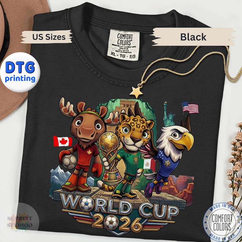 May include: A black t-shirt featuring a graphic of a cartoon moose, jaguar, and bald eagle standing on a platform with a soccer ball and trophy. The text reads World Cup 2026. The shirt includes a Comfort Colors label in size XL and DTG printing.