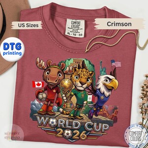 May include: A crimson t-shirt featuring a graphic of a cartoon moose, jaguar, and bald eagle standing on a platform with a soccer trophy. The text reads WORLD CUP 2026. The design includes flags of Canada, Mexico, and the United States, plus the Statue of Liberty.