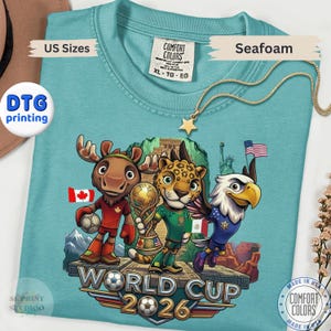 May include: A seafoam green t-shirt featuring a graphic of a cartoon moose, jaguar, and bald eagle holding a soccer ball and trophy. The design includes the text World Cup 2026, flags of Canada, Mexico, and the United States, and a pyramid background.