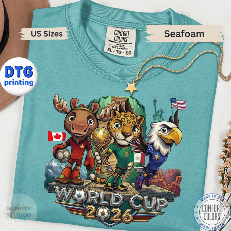 May include: A seafoam green t-shirt featuring a graphic of a cartoon moose, jaguar, and bald eagle holding a soccer ball and trophy. The design includes the text World Cup 2026, flags of Canada, Mexico, and the United States, and a pyramid background.