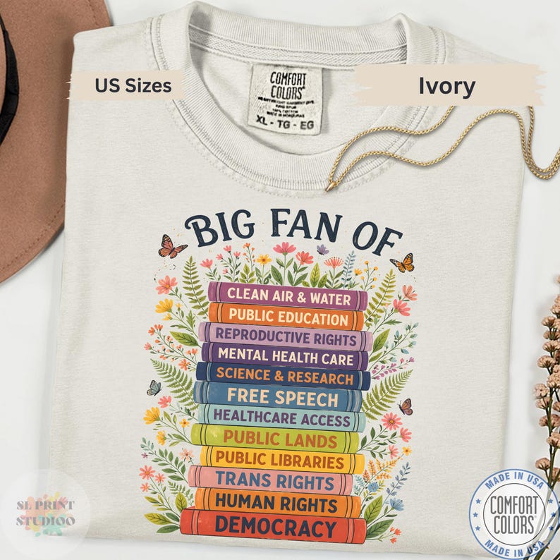 Ivory t-shirt featuring “Big Fan Of” text above colorful stacked books labeled democracy, human rights, public libraries, free speech, science, education, and healthcare access with floral details.