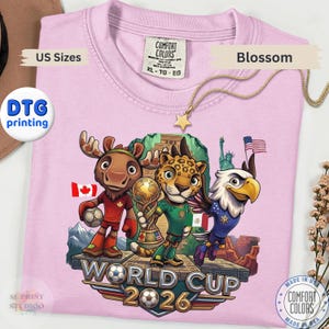 May include: A light pink t-shirt featuring a graphic of a cartoon moose, jaguar, and bald eagle standing on a stone platform. The text reads World Cup 2026. The moose holds a soccer ball, the jaguar holds a trophy, and the eagle is flying.