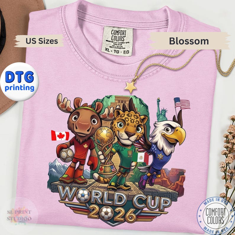 May include: A light pink t-shirt featuring a graphic of a cartoon moose, jaguar, and bald eagle standing on a stone platform. The text reads World Cup 2026. The moose holds a soccer ball, the jaguar holds a trophy, and the eagle is flying.