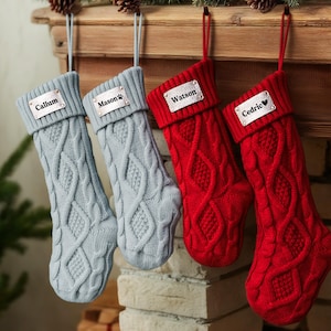 May include: Four cable-knit Christmas stockings in shades of grey and red hang from a wooden mantelpiece. Each stocking has a personalised tag with a name: Callum, Mason, Watson, and Cedric. The stockings are suspended by matching loops.