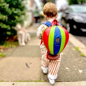 Könnte beinhalten: Ein Kinderrucksack in Form eines Heißluftballons mit roten, grünen, blauen und gelben Paneelen. Der Rucksack hat ein kleines gesticktes Heißluftballon-Design. Das Kind geht auf einem Bürgersteig, trägt gestreifte Hosen und weiße Schuhe.