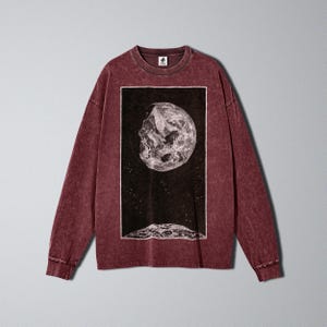 May include: A maroon long-sleeve shirt featuring a black and white graphic of a planet and stars. The graphic is rectangular and centered on the front of the shirt. The shirt has a crew neck and long sleeves.