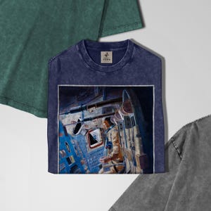 May include: A navy blue t-shirt with a vintage-style graphic of an astronaut inside a spacecraft. The graphic is framed in white. The shirt has a crew neck and a small tag near the neckline. Other shirts in green and gray are partially visible.