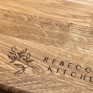 Può includere: Un primo piano di un tagliere di legno con il testo "Rebecca's Kitchen" e un piccolo disegno floreale inciso sulla superficie.