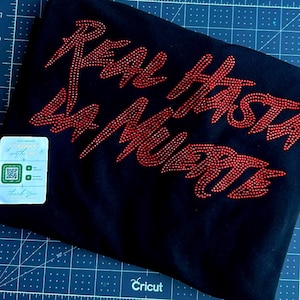 May include: Black t-shirt with the phrase "REAL HASTA LA MUERTE" in red rhinestones. The shirt has a tag with a QR code and the words "Connect with us". The shirt is on a blue cutting mat.