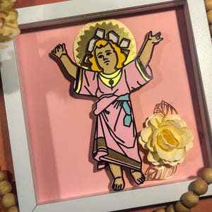May include: A framed artwork featuring a stylized depiction of a child figure with outstretched arms, wearing a pink robe with gold accents. The artwork is set against a pink background, with a gold halo and a paper flower in the corner.