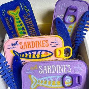 May include: A collection of colorful, rectangular containers labeled "not SARDINES" with fish illustrations. The containers are blue, pink, and purple, with yellow and blue accents. The text "SKINLESS + BONELESS" is also visible.
