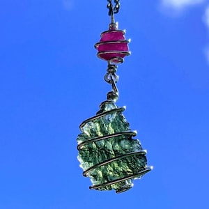 May include: A silver wire-wrapped pendant featuring a green, rough-cut gemstone and a pink gemstone. The pendant is hanging against a blue sky.