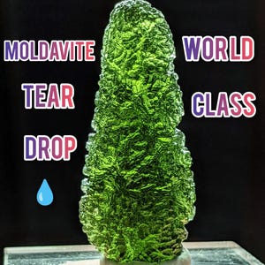 Op de afbeelding: Een levendige groene Moldaviet edelsteen, traanvormig, met een textuur. De afbeelding bevat de woorden "Moldavite Tear Drop World Class". De edelsteen wordt tegen een donkere achtergrond getoond.