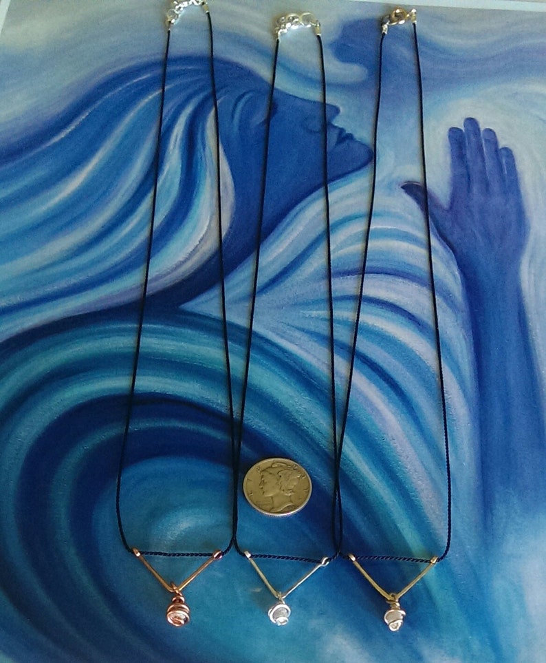 May include: Three necklaces with different colored pendants on black cords. The pendants are a rose gold, silver, and gold color. The necklaces are displayed on a blue and white background.