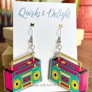 May include: A pair of retro-style earrings featuring a vibrant boombox design. The earrings showcase a teal body with pink and yellow accents, and silver-colored ear wires. The card behind the earrings reads "Quirks & Delight."