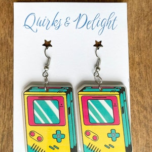 May include: A pair of dangle earrings featuring a yellow, pink, and blue retro handheld game console design. The earrings are silver and have a star-shaped hook.