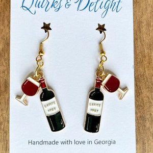 Könnte beinhalten: Ein Paar goldene Ohrringe mit einem roten Weinglas und einem Flaschenanhänger. Die Flasche ist mit "Lafite 1982" beschriftet. Die Ohrringe sind handgefertigt mit Liebe in Georgien.