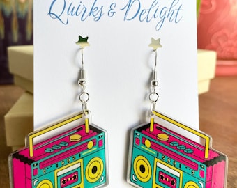 Puede incluir: Un par de pendientes de estilo retro con un diseño de radiocasete. Los pendientes presentan un cuerpo turquesa con detalles rosas y amarillos, y ganchos plateados. La tarjeta detrás de los pendientes dice "Quirks & Delight."