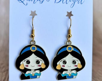 May include: A pair of gold-toned dangle earrings featuring a cartoon character design. The earrings depict a character with black hair, a blue headband, and a blue outfit. The card behind the earrings reads "Quirks & Delight" and "Handmade with love in Georgia."